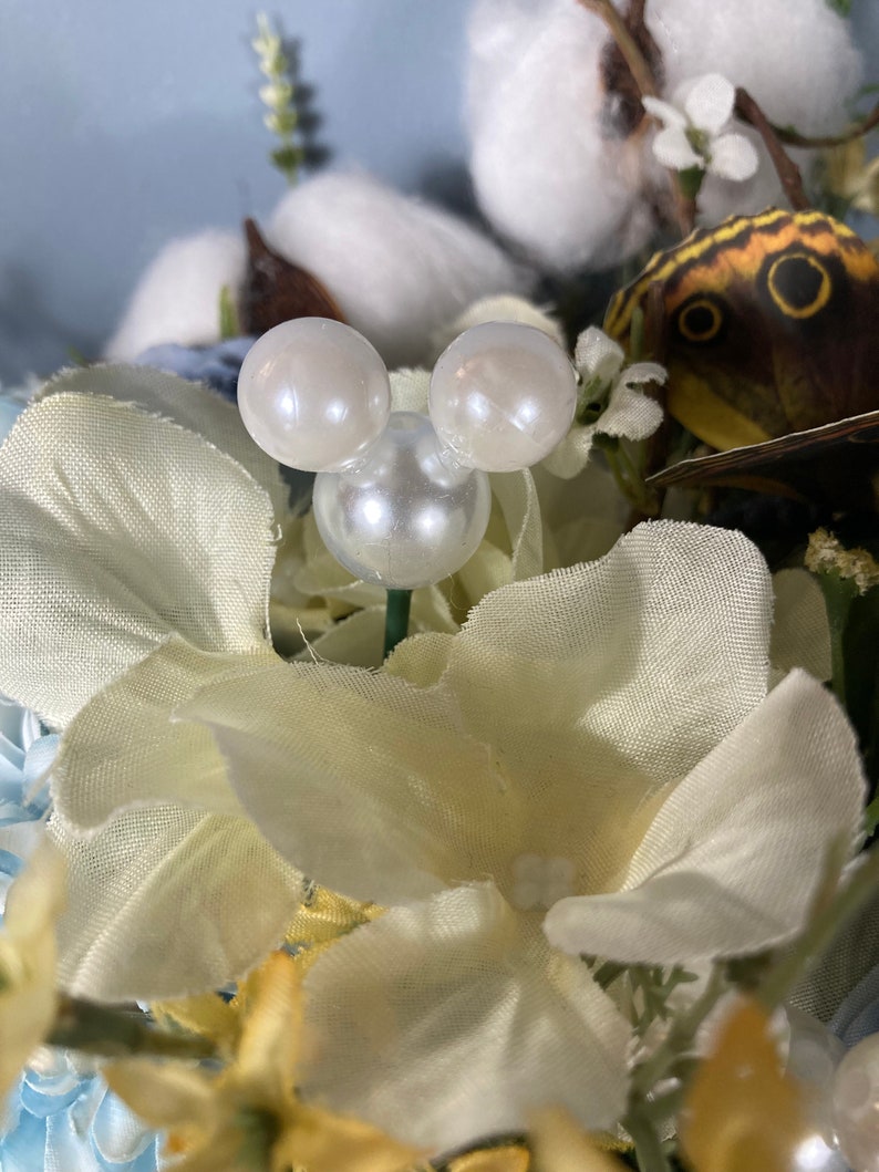 May include: A white fabric flower with a pearl Mickey Mouse head in the center. The flower is surrounded by other flowers and greenery.