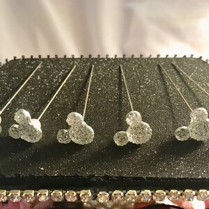 May include: Six silver-toned Mickey Mouse head pins with sparkling, glittery details are displayed on a black, glitter-covered surface. The pins are arranged in a row, with the Mickey Mouse heads facing upwards. The pins are likely decorative.