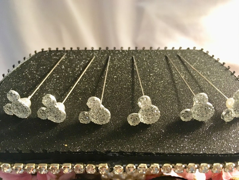 May include: Six silver-toned Mickey Mouse head pins with sparkling, glittery details are displayed on a black, glitter-covered surface. The pins are arranged in a row, with the Mickey Mouse heads facing upwards. The pins are likely decorative.