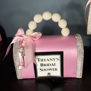 May include: Pink satin bridal shower gift box with a pearl handle, a silver key, and a white label that says "Tiffany's Bridal Shower".