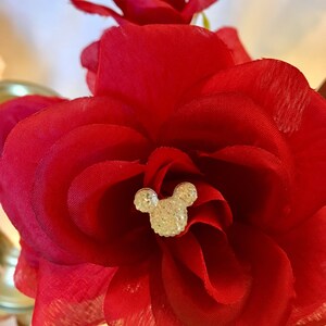 May include: Close-up of a vibrant red fabric rose with layered petals. A sparkling, crystal-studded Mickey Mouse head is centered in the flower. The image showcases a decorative floral arrangement, possibly for crafts or special events.