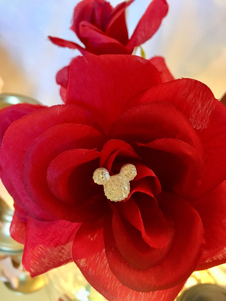 May include: Close-up of a vibrant red fabric rose with layered petals. A sparkling, crystal-studded Mickey Mouse head is centered in the flower. The image showcases a decorative floral arrangement, possibly for crafts or special events.