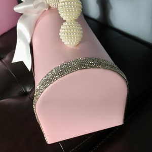 May include: A pink, decorative box with a white satin ribbon and a pearl trim. The box has a rhinestone trim around the top edge.