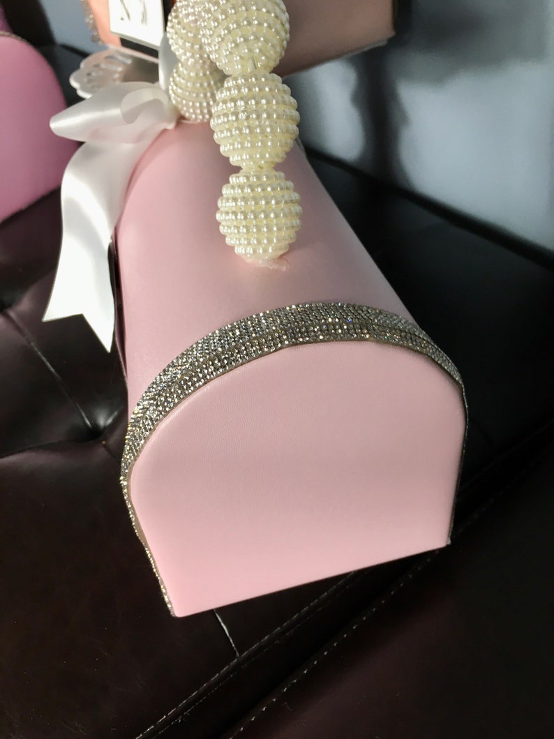 May include: A pink, decorative box with a white satin ribbon and a pearl trim. The box has a rhinestone trim around the top edge.