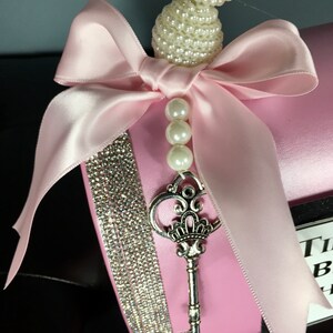 May include: Pink gift box with a large pink satin bow, a silver key, and a string of white pearls. The box is decorated with a strip of rhinestones.
