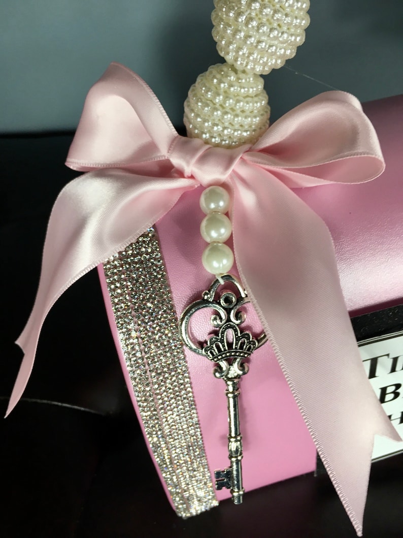 May include: Pink gift box with a large pink satin bow, a silver key, and a string of white pearls. The box is decorated with a strip of rhinestones.