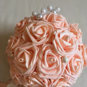 May include: A peach-colored bouquet of artificial roses with pearl accents. The bouquet is tied with a white and peach ribbon.