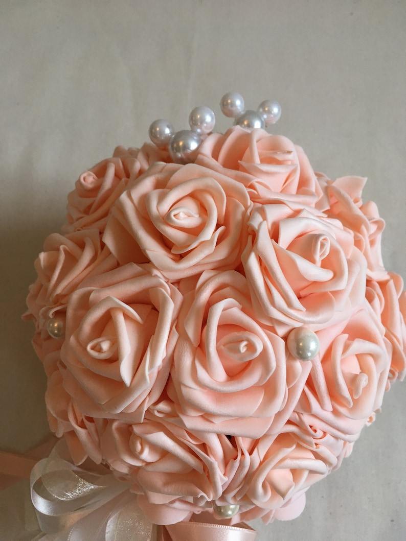 May include: A peach-colored bouquet of artificial roses with pearl accents. The bouquet is tied with a white and peach ribbon.