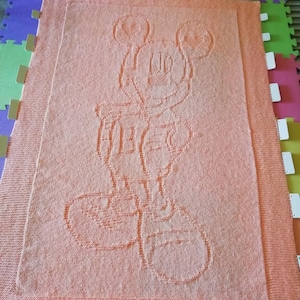 May include: A peach-colored knitted blanket with a white outline of Mickey Mouse in the center.