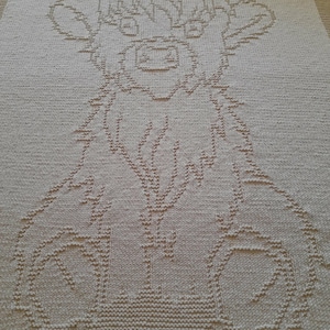 Può includere: Una coperta beige lavorata a maglia con un disegno di mucca delle Highlands. La mucca è raffigurata in uno stile semplice, con corna, occhi e un corpo soffice. La coperta è rettangolare e sembra fatta a mano.