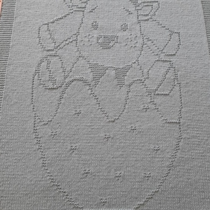 May include: A knitted blanket in a soft, muted teal color. The blanket features a detailed pattern of a cartoon reindeer inside an egg. The reindeer has large antlers and a smiling face. The blanket is a cozy, handmade item.
