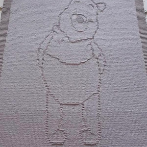 Könnte beinhalten: Eine hellgraue Strickdecke mit einem Winnie-the-Pooh-Design. Die Figur ist in einem dunkleren Grauton umrissen, mit Details wie Gesicht, Armen und Beinen. Die Decke hat einen einfachen Rand.