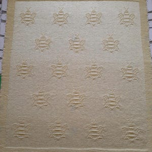 May include: A cream-coloured knitted blanket with a repeating pattern of bees. The blanket has a textured appearance with a light yellow border. The bee pattern is evenly spaced across the surface, creating a cosy and decorative textile.