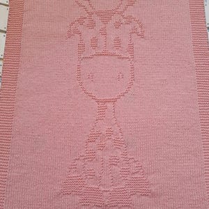 Könnte beinhalten: Eine rosa gestrickte Babydecke mit einem Giraffenmuster. Die Giraffe befindet sich in der Mitte der Decke und ist mit einem Reliefmuster gestrickt.