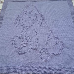 Puede incluir: Una manta de bebé de punto de color lavanda con un contorno blanco de un personaje de dibujos animados, posiblemente Eeyore de Winnie the Pooh.