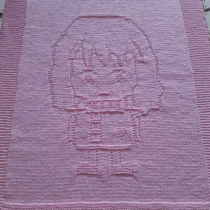 Puede incluir: Una manta de bebé de punto rosa con un diseño de cara de niña en el centro.