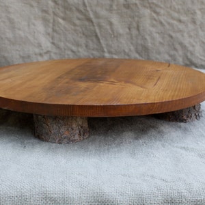 May include: A round, wooden serving tray with a natural finish. The tray is supported by three short, cylindrical legs made from tree branches. The wood has a warm, brown tone, and the tray is suitable for displaying food or decorative items.