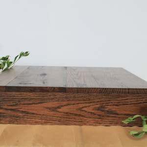 May include: A rectangular wooden box with a dark brown finish. The box is made of planks of wood and has a natural wood grain pattern. Green leafy sprigs are placed on the side and top of the box.