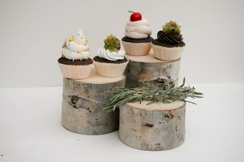 Puede incluir: Cuatro cupcakes decorados en un soporte de madera de varios niveles. Los cupcakes est&aacute;n cubiertos con glaseado blanco y varias decoraciones, incluyendo chispas, una cereza y una hoja verde. El soporte est&aacute; hecho de madera natural con un acabado r&uacute;stico.