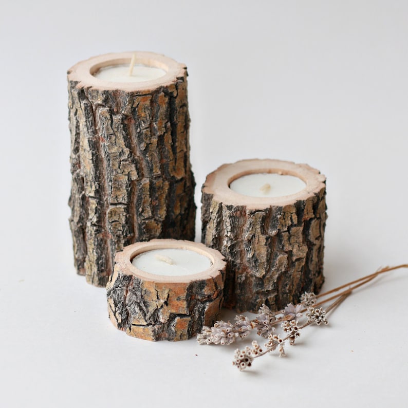 Pu&ograve; includere: Tre portacandele in legno realizzati con corteccia d'albero con candele bianche all'interno. I portacandele sono disposti a forma di piramide su una superficie bianca.