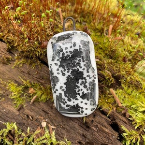 Può includere: Un ciondolo in vetro bianco con un design geometrico bianco e nero. Il ciondolo ha un anello d'argento in cima per appenderlo.