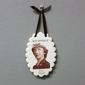 May include: White ceramic ornament with a scalloped edge, featuring a black and white photo of Grace Hopper wearing a hat with the text "BUG ROYALTY" above the photo and "Grace Hopper" below the photo.