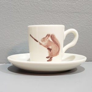 May include: A white ceramic coffee cup with a brown squirrel holding a rifle printed on the side. The cup is sitting on a white saucer.
