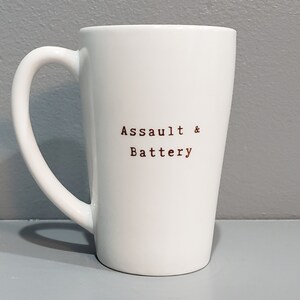以下が含まれることがあります： 黒い取っ手を持つ白いセラミックマグカップ。マグカップには、茶色のフォントで「Assault & Battery」という文字が前面に印刷されています。