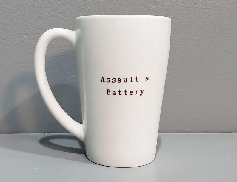 以下が含まれることがあります： 黒い取っ手を持つ白いセラミックマグカップ。マグカップには、茶色のフォントで「Assault & Battery」という文字が前面に印刷されています。