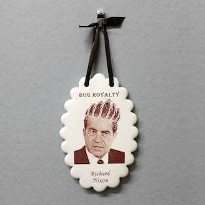 May include: A white ceramic ornament with a scalloped edge, featuring a portrait of Richard Nixon with a crown of bugs. The ornament reads "Bug Royalty" and "Richard Nixon".