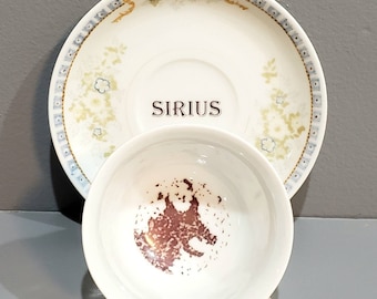 May include: A white teacup and saucer set with a floral design. The saucer has a blue and gold border and the text "SIRIUS" printed on the bottom. The teacup has a brown stain on the inside.