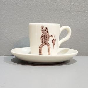 May include: A white ceramic espresso cup and saucer set. The cup features a brown illustration of a frog holding an umbrella. The cup has a curved handle and sits on a matching saucer. The set is designed for serving coffee or espresso.
