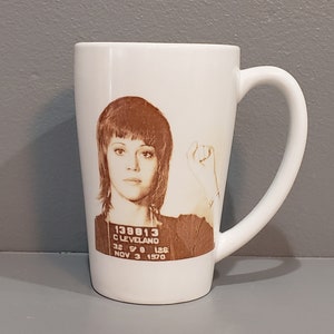 以下が含まれることがあります： 茶色の短い髪の女性が描かれた白いセラミック製のマグカップ。画像は、女性の顔写真で、その下に「138813 CLEVELAND 32 5'0 125 NOV 3 1970」というテキストが印刷されています。