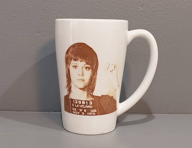 以下が含まれることがあります： 茶色の短い髪の女性が描かれた白いセラミック製のマグカップ。画像は、女性の顔写真で、その下に「138813 CLEVELAND 32 5'0 125 NOV 3 1970」というテキストが印刷されています。