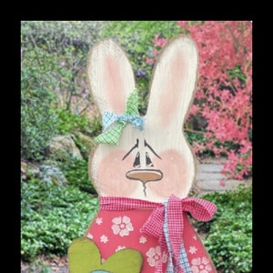 以下が含まれることがあります： ピンクと白のストライプの脚、白い花がついたピンクのドレス、緑の花、青と白のリボンをつけた木製のウサギの置物。ウサギは木製の台座の上に立っています。