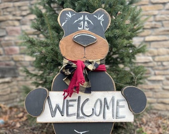 May include: Wooden black bear cutout holding a sign that says "WELCOME". The bear is wearing a red and black plaid scarf.