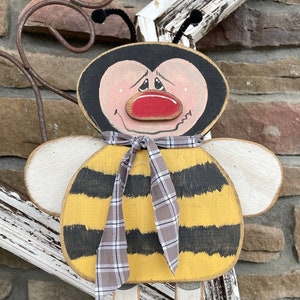 May include: A wooden bee figurine with a yellow and black striped body, white wings, and a red nose. The bee is wearing a plaid scarf and has black painted legs and feet. The bee is standing on a wooden base.