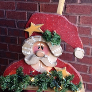 May include: Wooden Santa Claus cutout with a red hat, white beard, and a red and white striped shirt. The cutout is decorated with green garland and white lights. The cutout is standing in front of a brick wall with two wooden signs that read "Christmas Blessings".