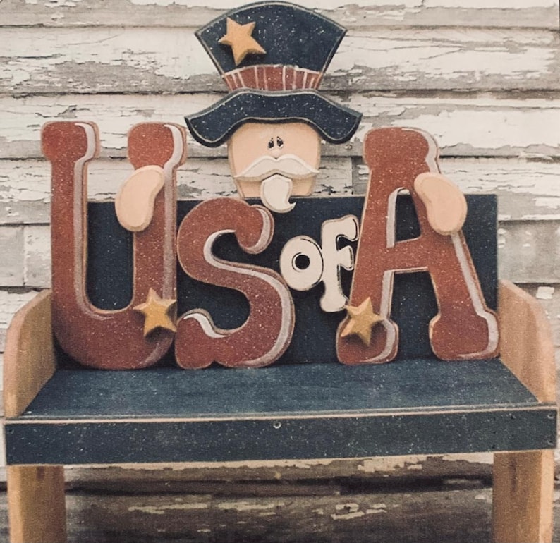 May include: A wooden bench with a patriotic theme. The bench is painted blue and has a wooden sign that says "US of A" in red letters with gold stars. A wooden Uncle Sam figure is sitting on top of the sign.