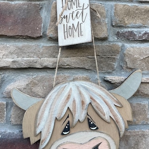 以下が含まれることがあります： ピンクの鼻と黒い目をした木製のハイランド牛の頭。牛は、「Home Sweet Home」と書かれた木製のタグが付いた麻ひもで吊り下げられています。