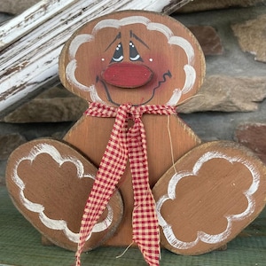 May include: A wooden gingerbread man figurine with a red nose, white eyes, and a red and white checkered ribbon around its neck. The figurine is painted brown and has white accents.