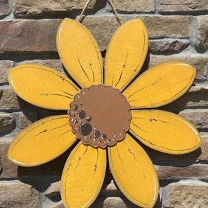 May include: A wooden sunflower decoration with yellow petals and a brown centre. The petals have a distressed look with black lines and white edges. A twine loop is attached for hanging.