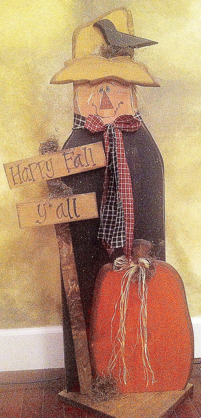 K&ouml;nnte beinhalten: Eine Holz-Vogelscheuche mit einem schwarzen Raben auf ihrem Hut, die ein Schild mit der Aufschrift "Happy Fall y'all" h&auml;lt. Die Vogelscheuche tr&auml;gt einen rot-schwarzen karierten Schal und h&auml;lt einen gro&szlig;en orangefarbenen K&uuml;rbis.