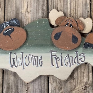May include: Wooden sign in the shape of a fish, painted with a brown moose design and the words "Welcome Friends". The moose has large brown eyes and antlers. The sign is painted in shades of green, brown, and white.
