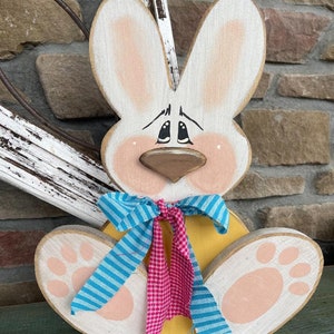 May include: A wooden Easter bunny with a yellow body, white ears, and pink feet. The bunny is wearing a blue and white striped bow and a pink and white checkered bow.