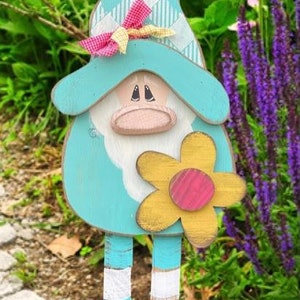 May include: A wooden gnome figurine with a blue and white checkered hat, a pink bow, and a yellow flower with a pink center. The gnome is standing on a wooden base with a blue and white striped pattern.