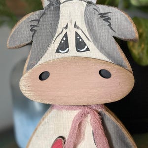 Peut inclure: Figurine de vache en bois avec une finition vieillie. La vache a un nez rose, une écharpe rose et un cœur rouge découpé. La tête est grise et blanche avec des yeux noirs. Un objet décoratif charmant et rustique.
