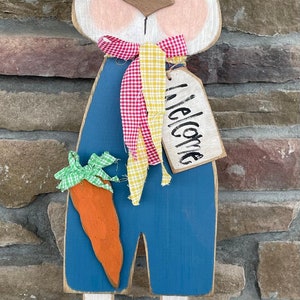 以下が含まれることがあります： 青い白のストライプの服を着た、ニンジンと赤と黄色のチェックのリボンをつけた木製のウサギの切り抜き。ウサギは「ようこそ」と書かれた看板を持っています。