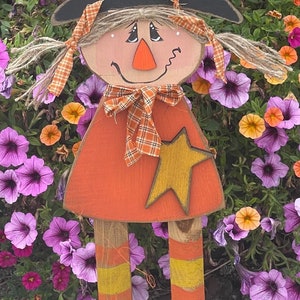 May include: A wooden witch figurine with a black hat, orange dress, and yellow striped legs. The witch has a friendly face with a black nose and brown eyes. The figurine is standing in front of a bed of purple and yellow flowers.