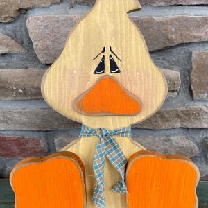 May include: A wooden duck figurine with a distressed paint finish. The duck is painted in shades of brown, orange, and blue. It has a large, round head with a painted face, and a small, tied bow around its neck.